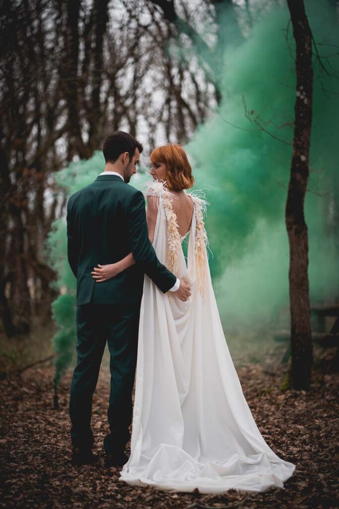 mariés vus de dos dans la foret, devant des fumigènes vert. ils se tiennent par la taille. La mariée porte une cape
