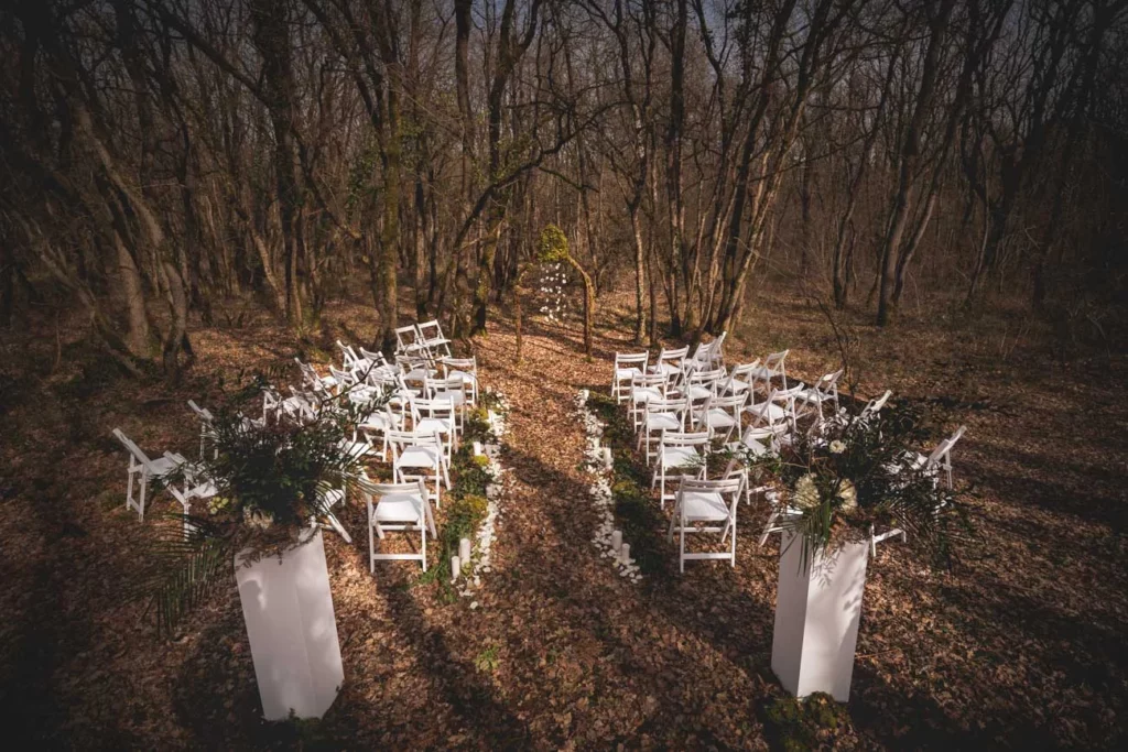 cérémonie de mariage en automne, chaises blanche disposées en arc de cercle, deux compositions florales naturelles à l’entrée. Scène  de jour en forêt