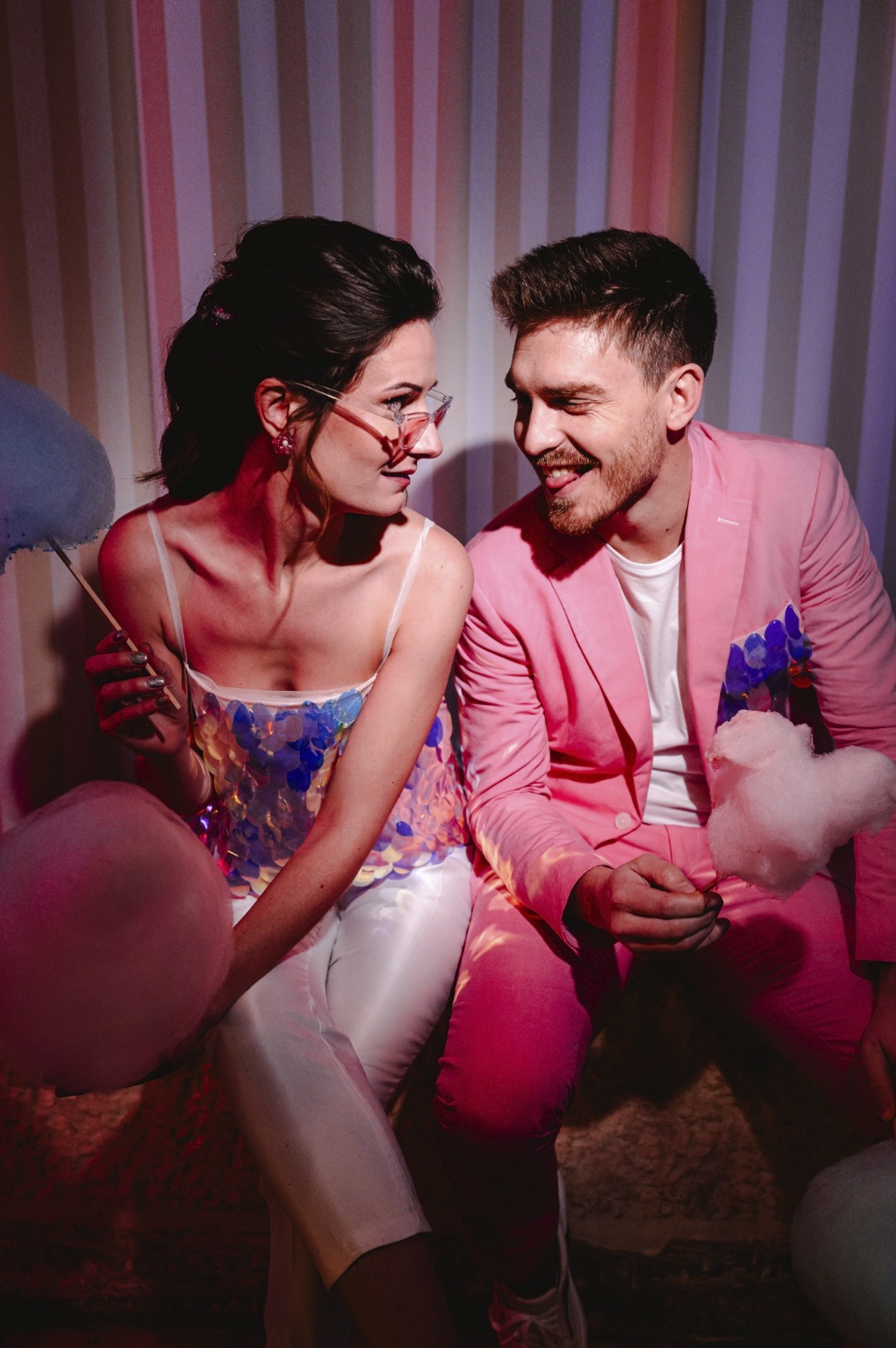 Un couple assis côte à côte, souriant et profitant d'un moment lors d'une fête. La femme tient un bâton de barbe à papa, et ils sont tous les deux habillés de manière vibrante et ludique, ce qui reflète l'atmosphère festive de la fête, avec un fond éclairé au néon dans des nuances de rose et de violet