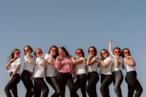 Groupe de femmes en tenue assortie, t-shirts blancs et pantalons noirs, portant des lunettes en forme de cœur rouges. Elles posent sur une plage sous un ciel bleu, entourant la future mariée vêtue d'un haut rose à motifs. Ambiance fun et complice pour un EVJF en plein air.